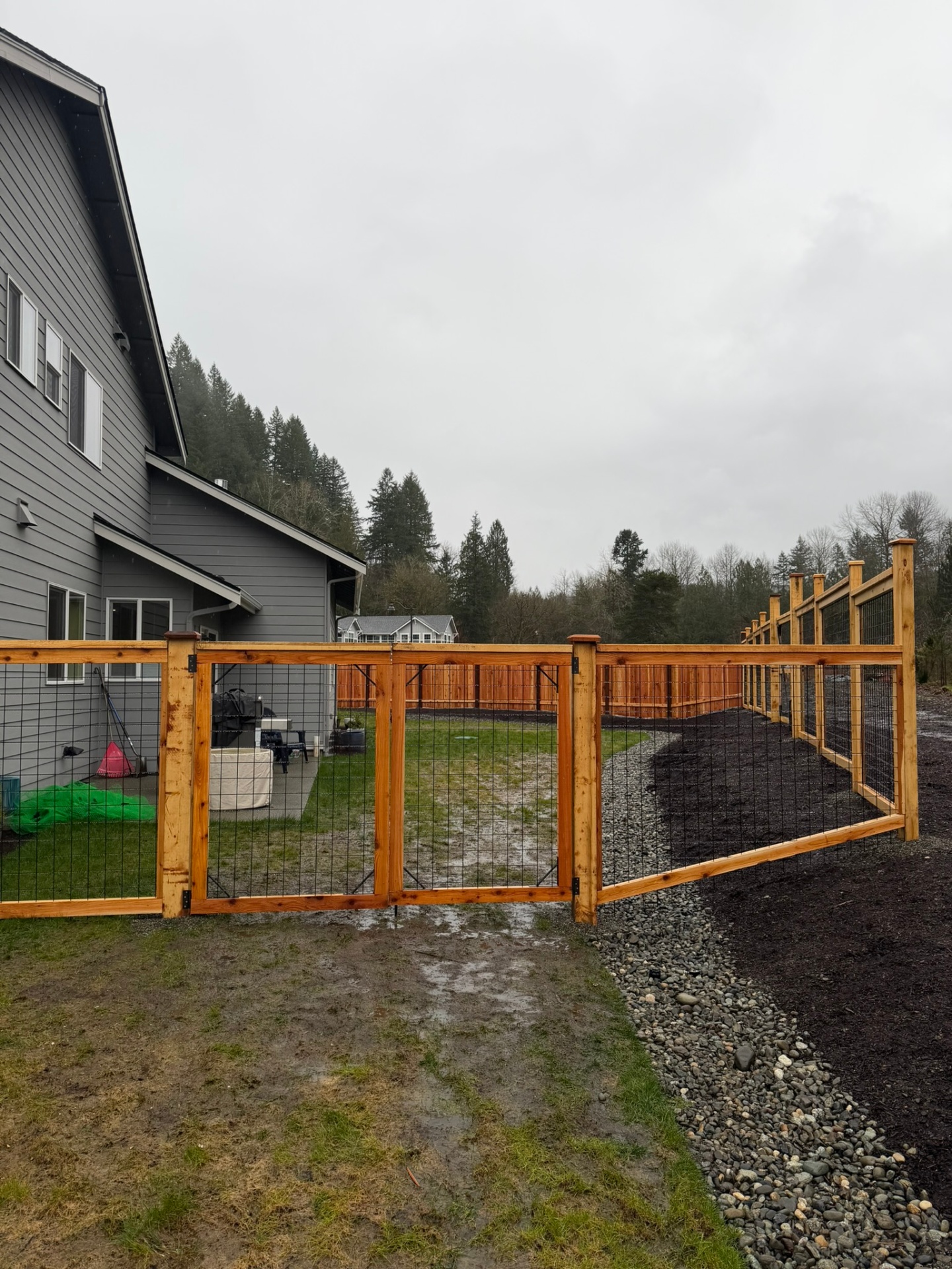 Cedar-framed hogwire dog-run fence with heavy double gate wrapping a modern gray farmhouse on a rural Puget Sound lot